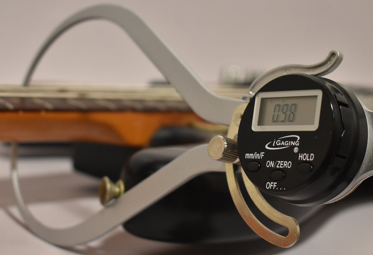 A digital caliper measures the string height above the frets on the bass's neck, showing "0.98" on its screen.