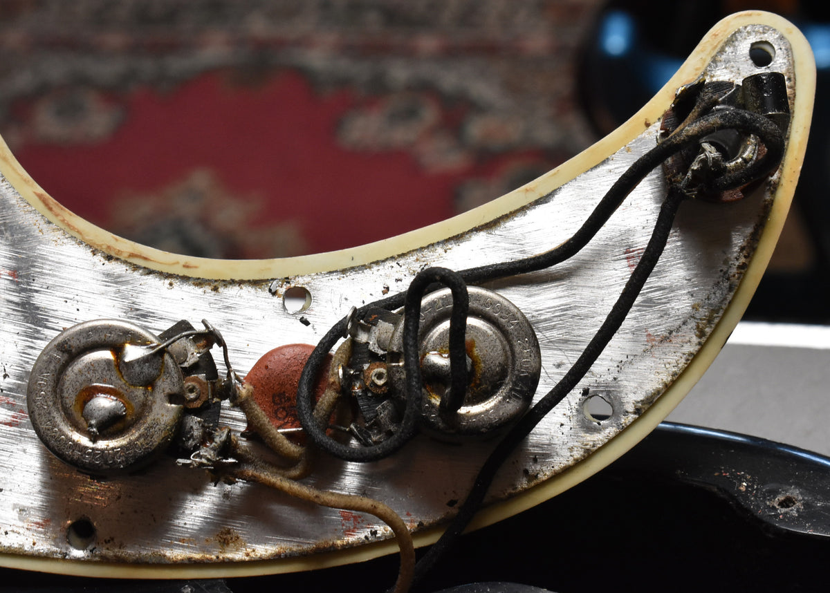 Close-up of the bass's control cavity, showing two tarnished pots, an orange capacitor, and cloth-wrapped wiring on a worn metal plate.