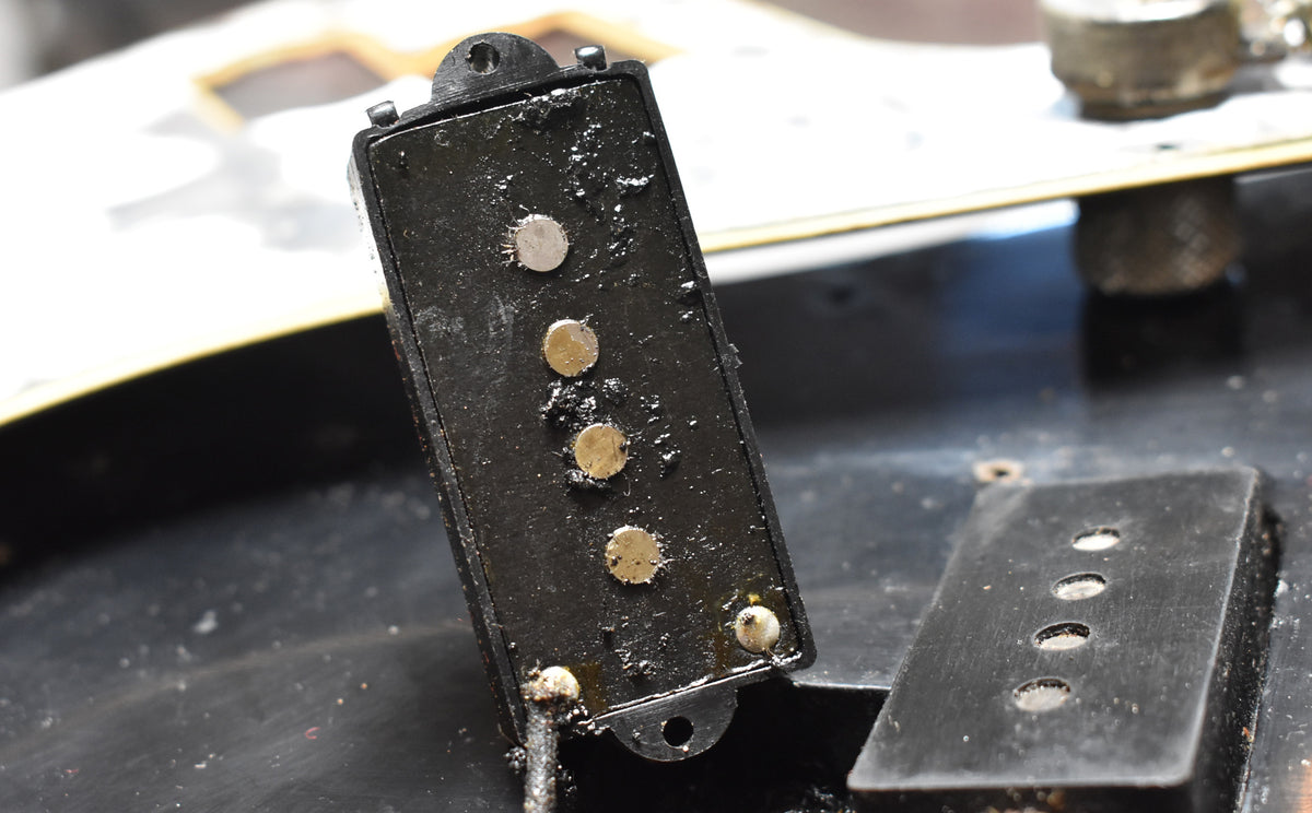 Close-up of a black, wax-potted split-coil pickup with four gold pole pieces and two silver ones, next to another pickup cover.