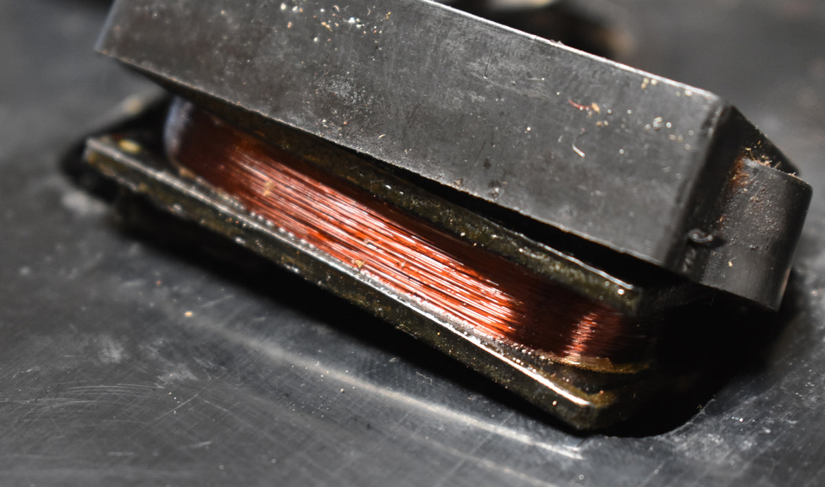 Close-up of a pickup with exposed copper wire windings, showing a dark, worn casing and some dust and debris.