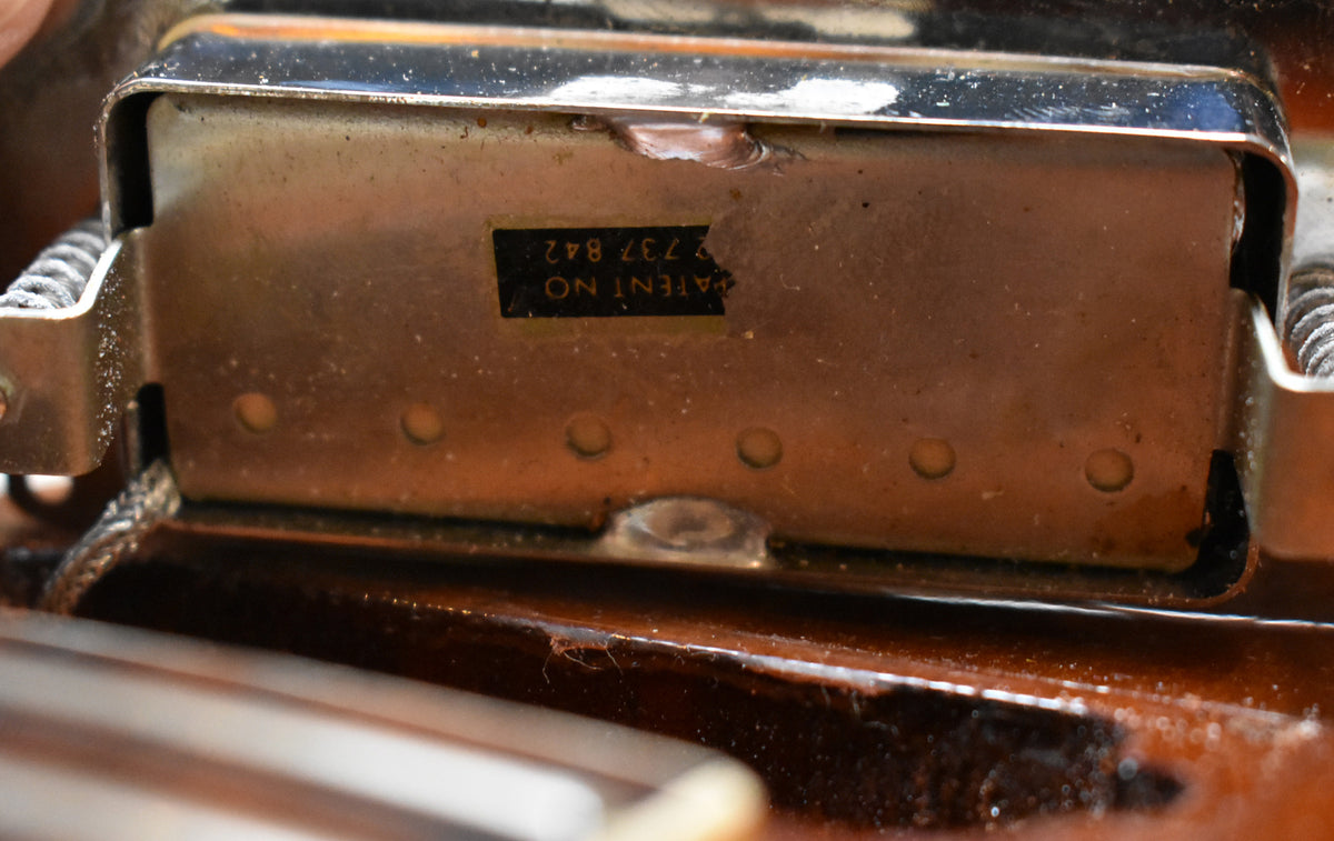 Close-up of a tarnished metal pickup cover with a partially torn black patent sticker, showing wear and dust on the surrounding sunburst finish.