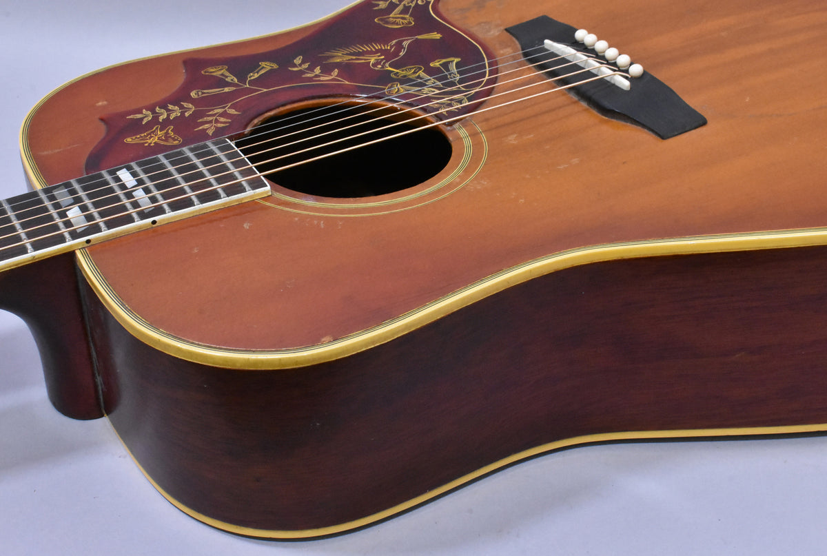 Upper body of a sunburst acoustic guitar, showing the dark red pickguard with gold hummingbird inlay, and some finish wear.