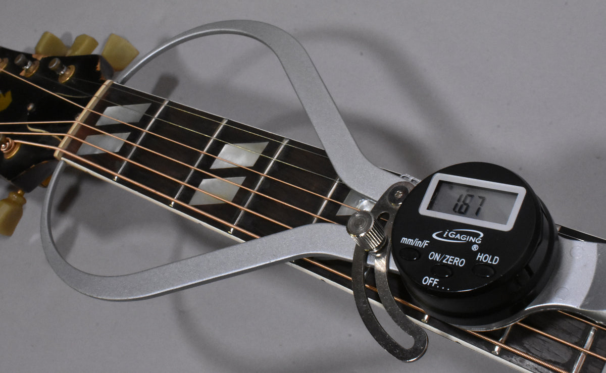 A digital string action gauge measures 1.67mm at the 12th fret of the guitar's dark fretboard with parallelogram inlays.