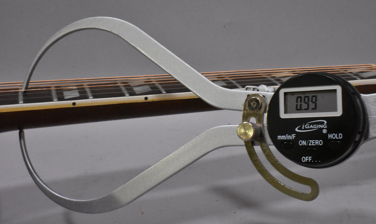 A digital caliper measures the string height at the 12th fret, showing 0.99mm, on the guitar's dark brown neck and fretboard.