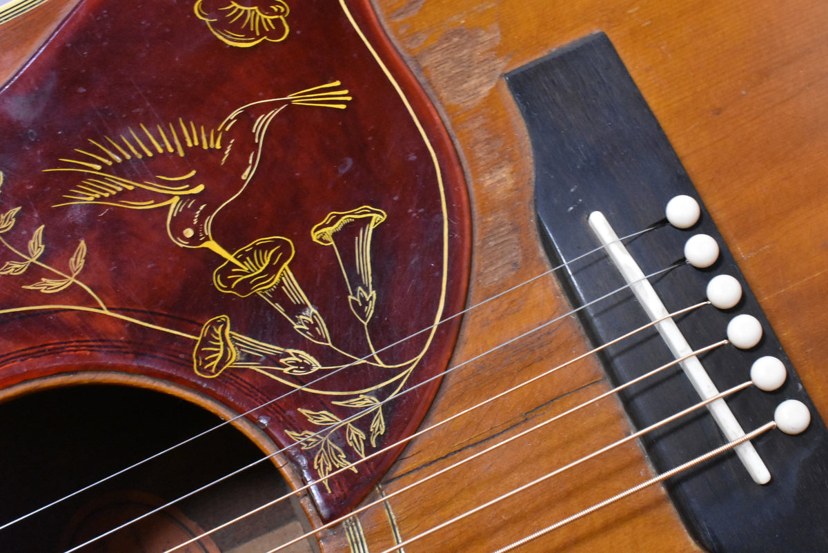 Close-up of the sunburst body, showing the pickguard with a hummingbird and flowers, and the bridge with six white pins.