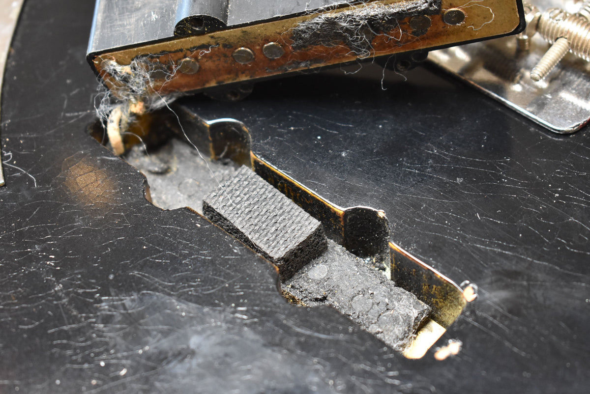 Close-up of the black body with heavy scratching and checking, revealing a cavity with foam padding and a rusty pickup base.