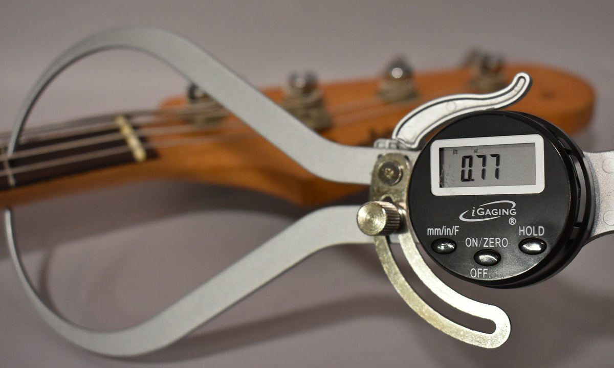 A digital caliper measures the bass's string height at the nut, displaying "0.77" on its screen, with the headstock blurred behind it.