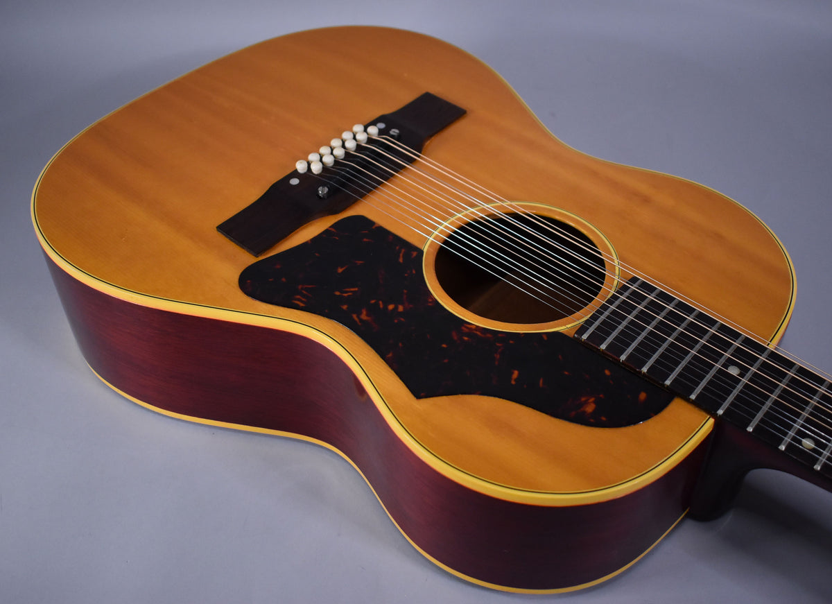The 12-string acoustic guitar's natural finish top and dark red sides are visible from above, with a tortoise pickguard.