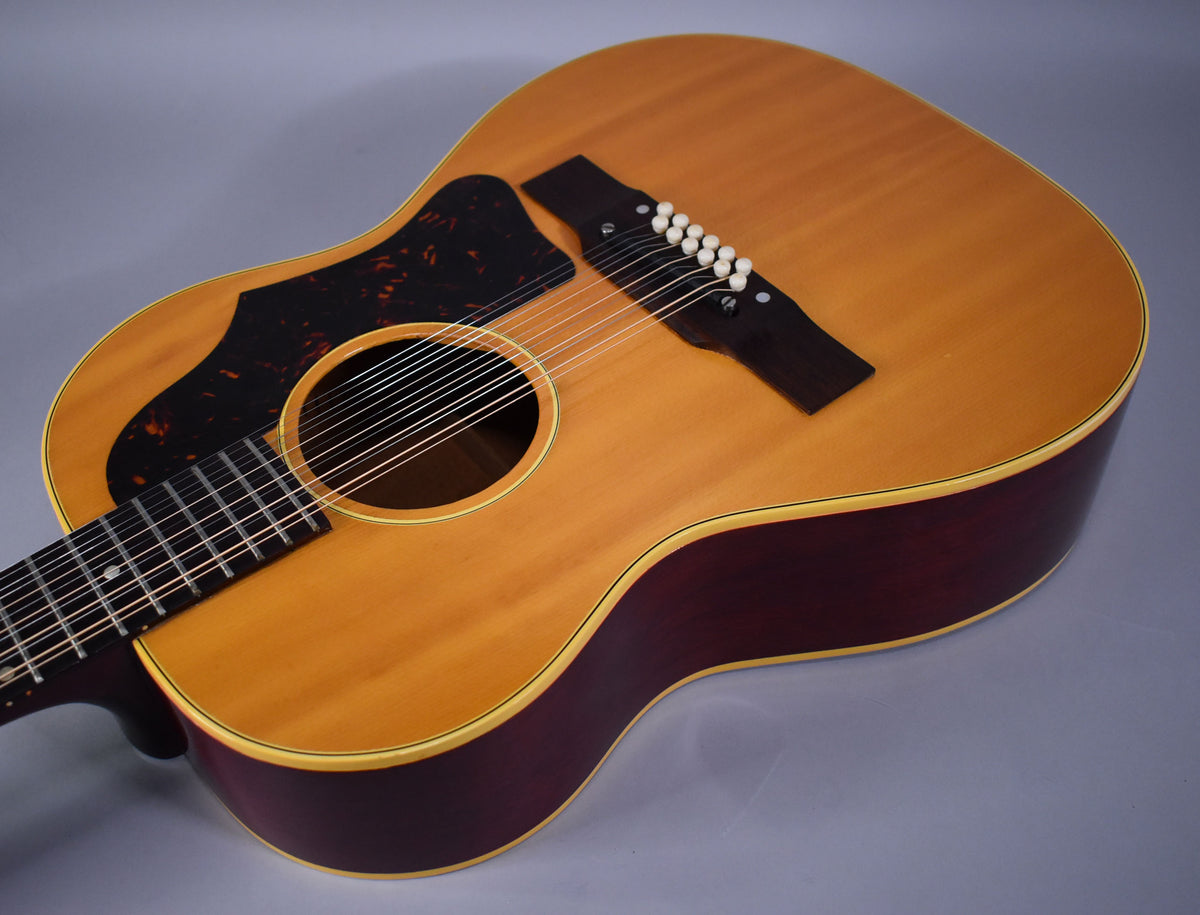 The natural finish 12-string acoustic guitar body is angled, showing its light brown top, dark red sides, and tortoiseshell pickguard.
