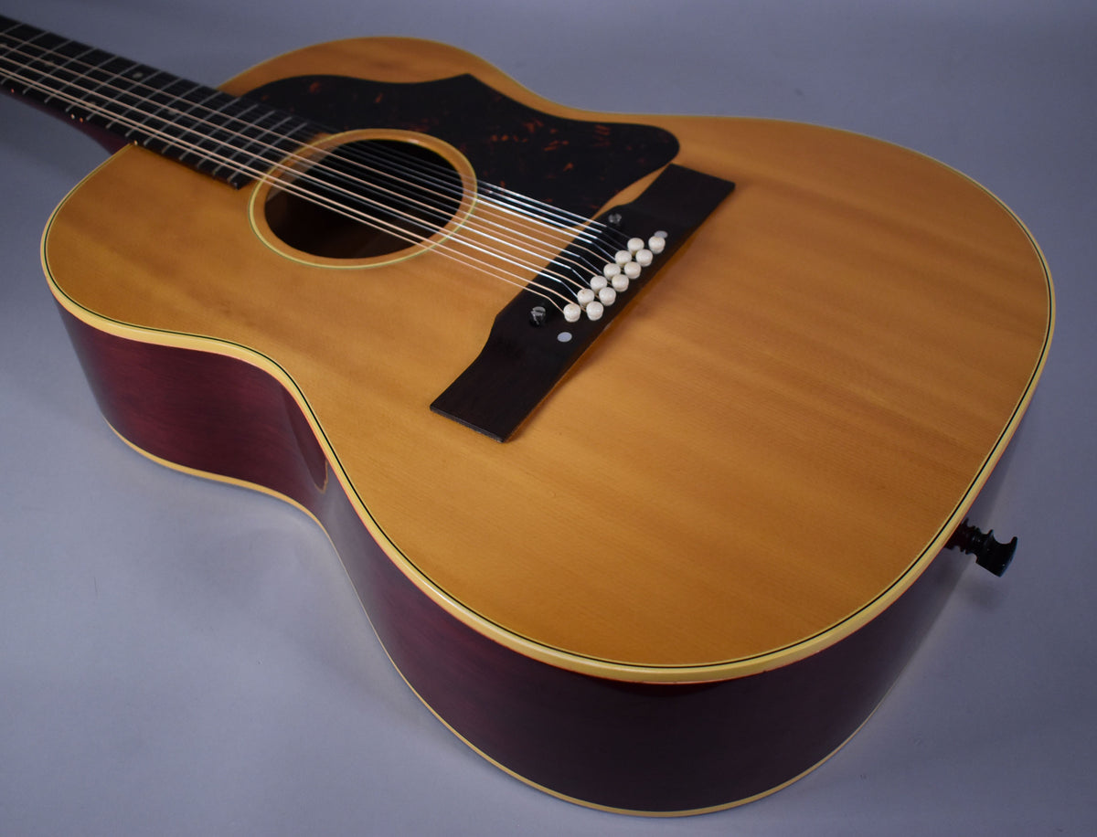 A natural finish 12-string acoustic guitar, angled from the neck, showing the light-colored top, dark sides, and black pickguard.