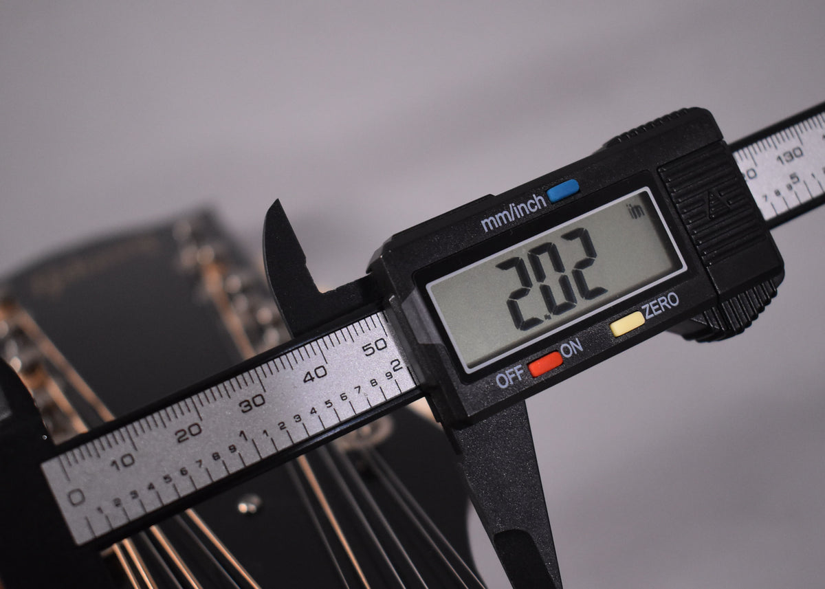 A digital caliper measures the guitar's nut width at 2.02 inches, with the headstock and strings blurred in the background.