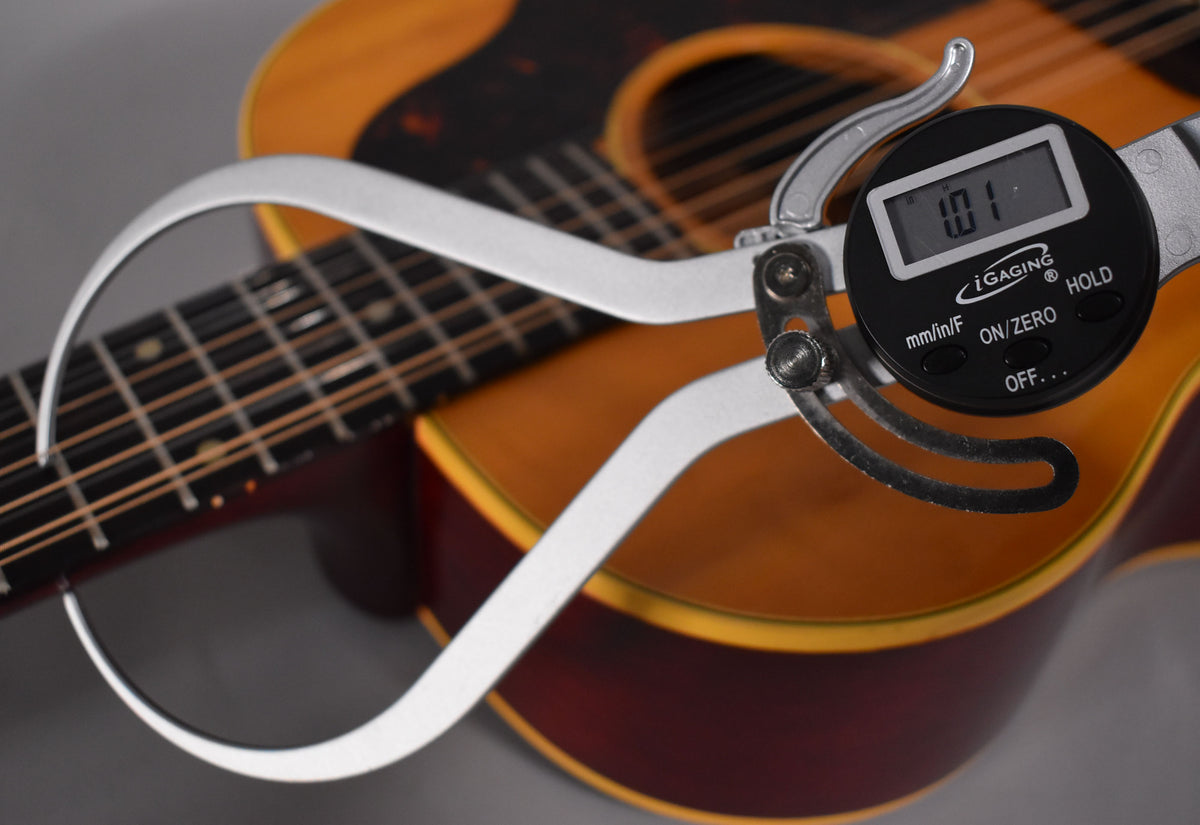 A digital caliper measures the string height at the 12th fret of the natural finish acoustic guitar.