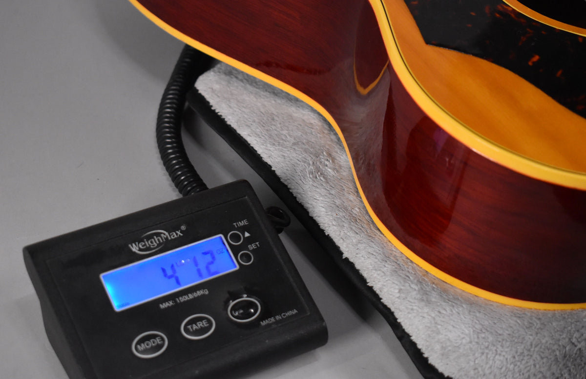 The guitar's dark red back and sides with yellow binding rest on a scale displaying "4.12" lbs, next to a soft gray lining.