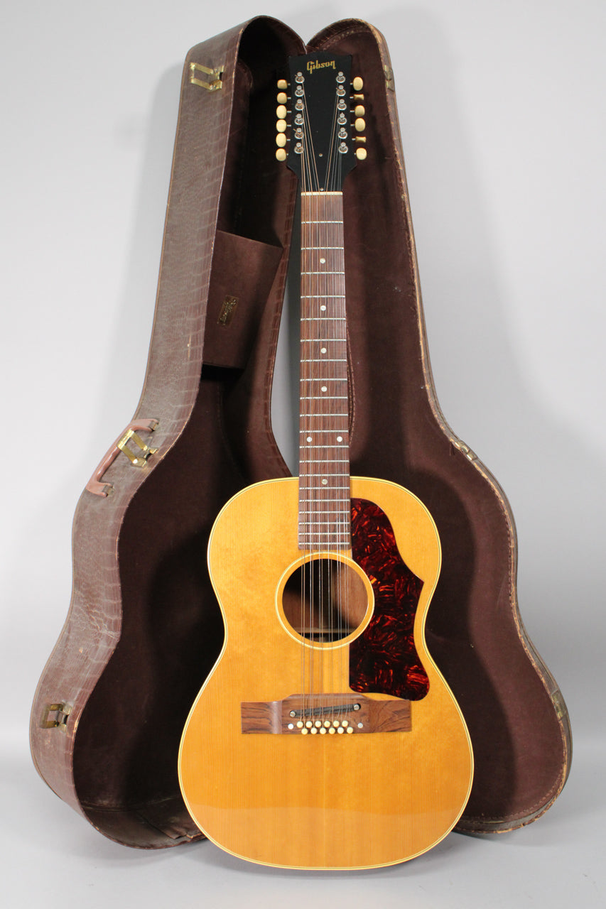 Full-length view of a natural finish 12-string acoustic guitar with a tortoiseshell pickguard, standing in its open brown hard case.