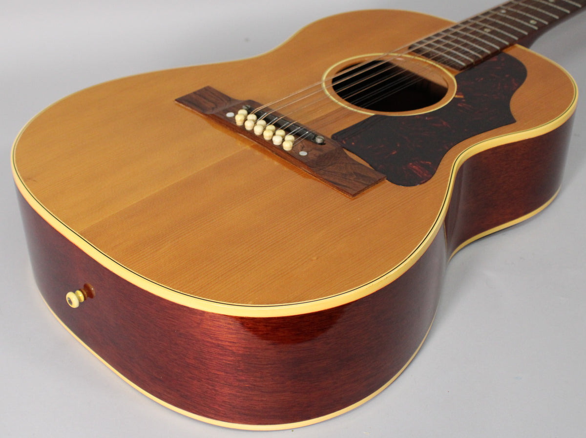 The natural finish 12-string acoustic guitar is angled from the lower bout, showing its dark red sides and tortoise pickguard.