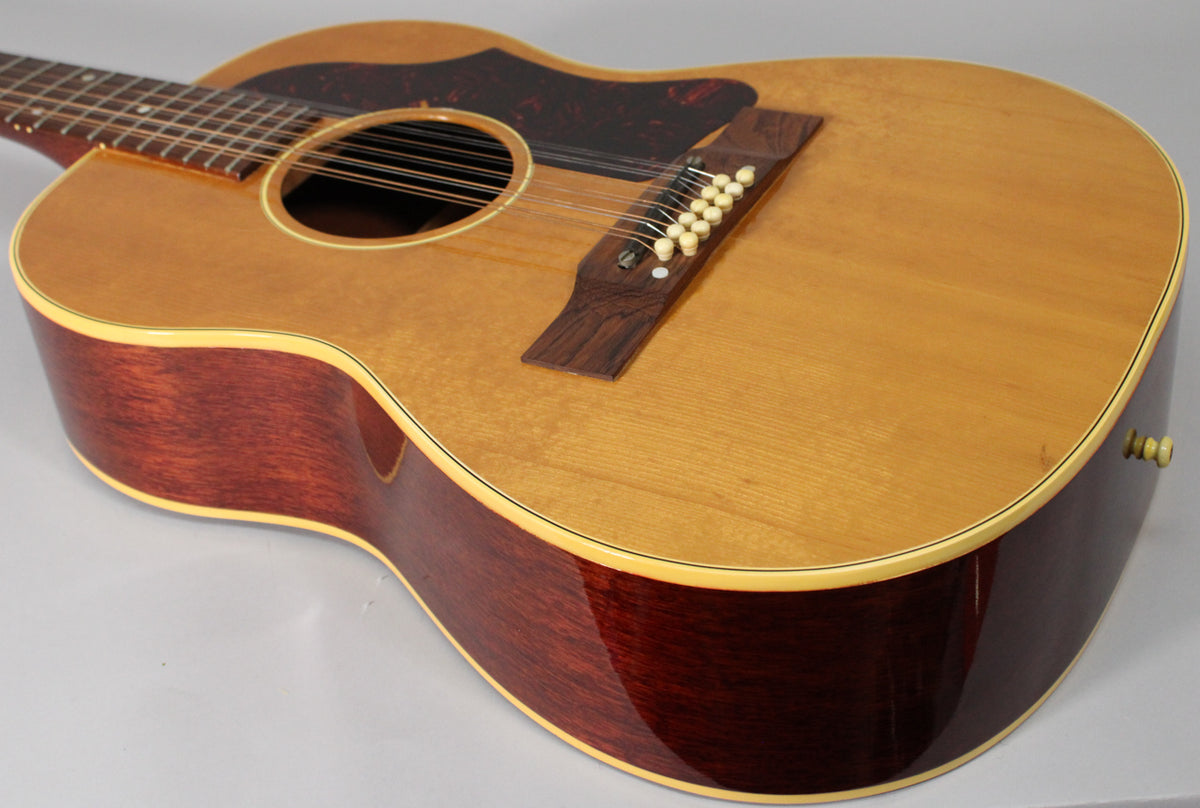 The upper body of a natural finish 12-string acoustic guitar, showing the dark pickguard, rosewood bridge, and cream binding.
