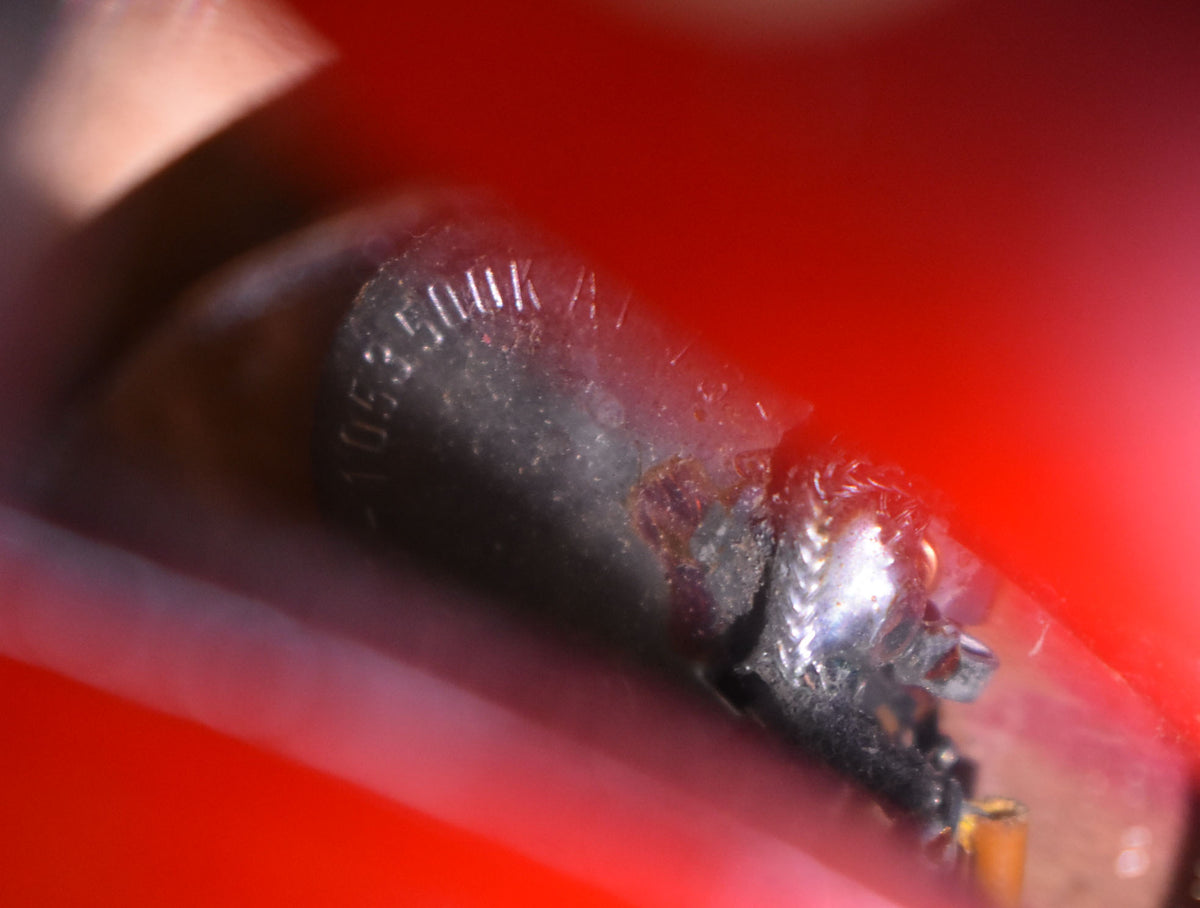 Extreme close-up of a dark, cylindrical component with faint white lettering, showing solder joints and wires against a blurred red background.