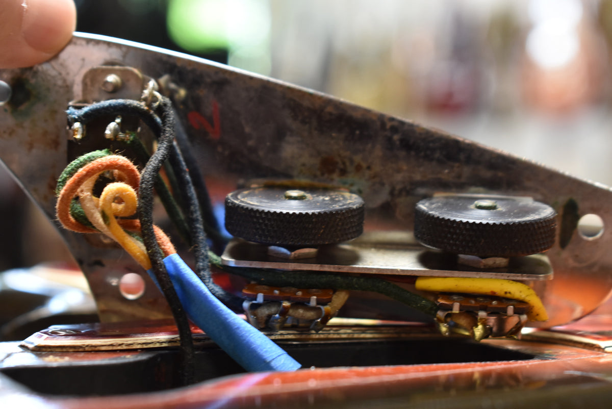 Close-up of the guitar's control cavity, revealing two knurled black roller knobs, aged wiring, and corroded metal shielding.