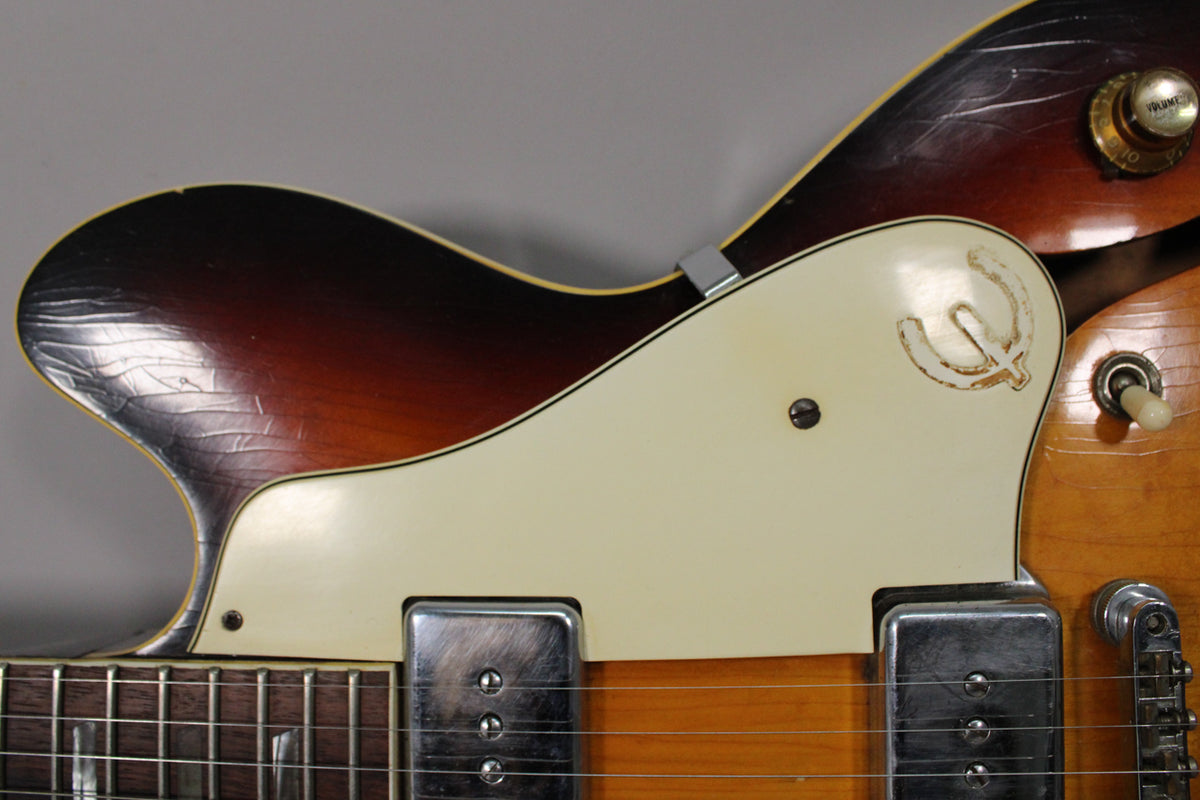 Close-up of the sunburst guitar's upper bout, showing a cream pickguard with a worn logo, two chrome pickups, and heavy finish checking.