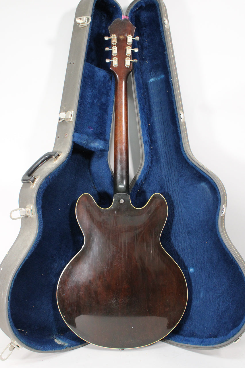 The guitar's dark sunburst back and neck are visible, showing finish wear and checking, resting in a grey hard case with blue lining.