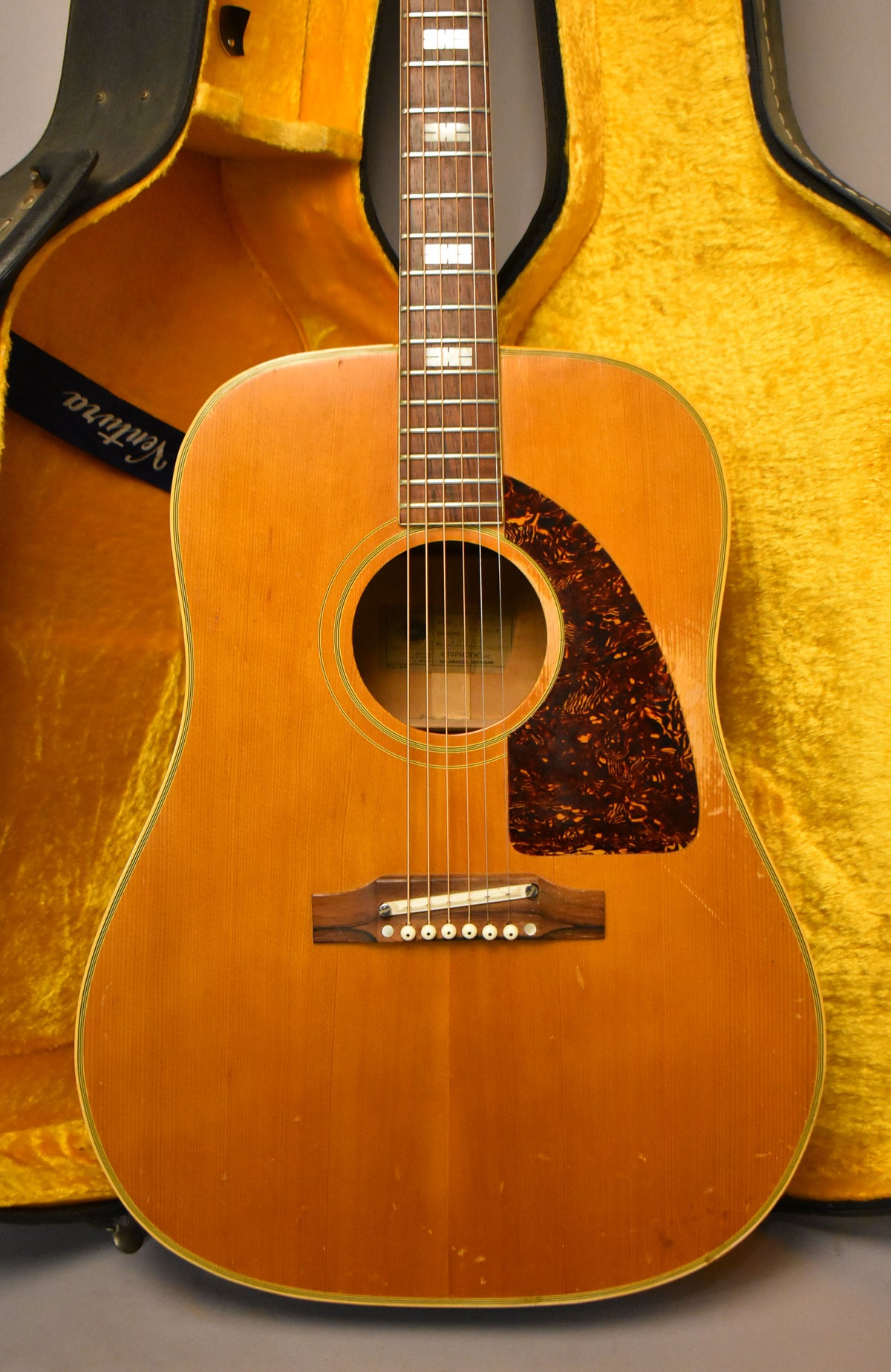 The natural finish acoustic guitar is angled slightly, showing its body, fretboard with block inlays, and a tortoiseshell pickguard.