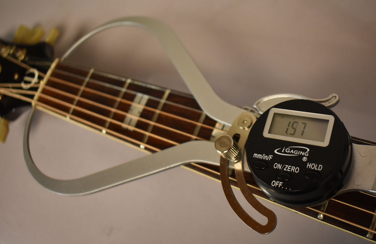 A digital caliper measures the string height at the 12th fret, showing 1.57mm, on the natural finish acoustic guitar.