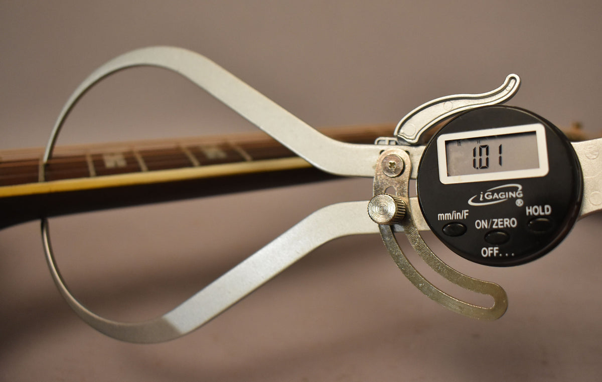 A digital caliper measures the guitar's neck relief at 0.01 inches, with the fretboard and frets visible in the background.