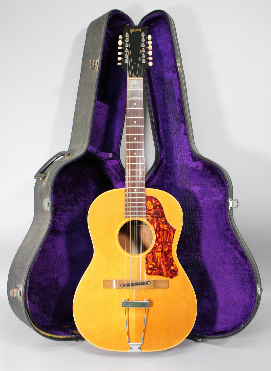 Full front view of a natural finish 12-string acoustic guitar with a tortoiseshell pickguard, standing in its open, purple-lined hard case.