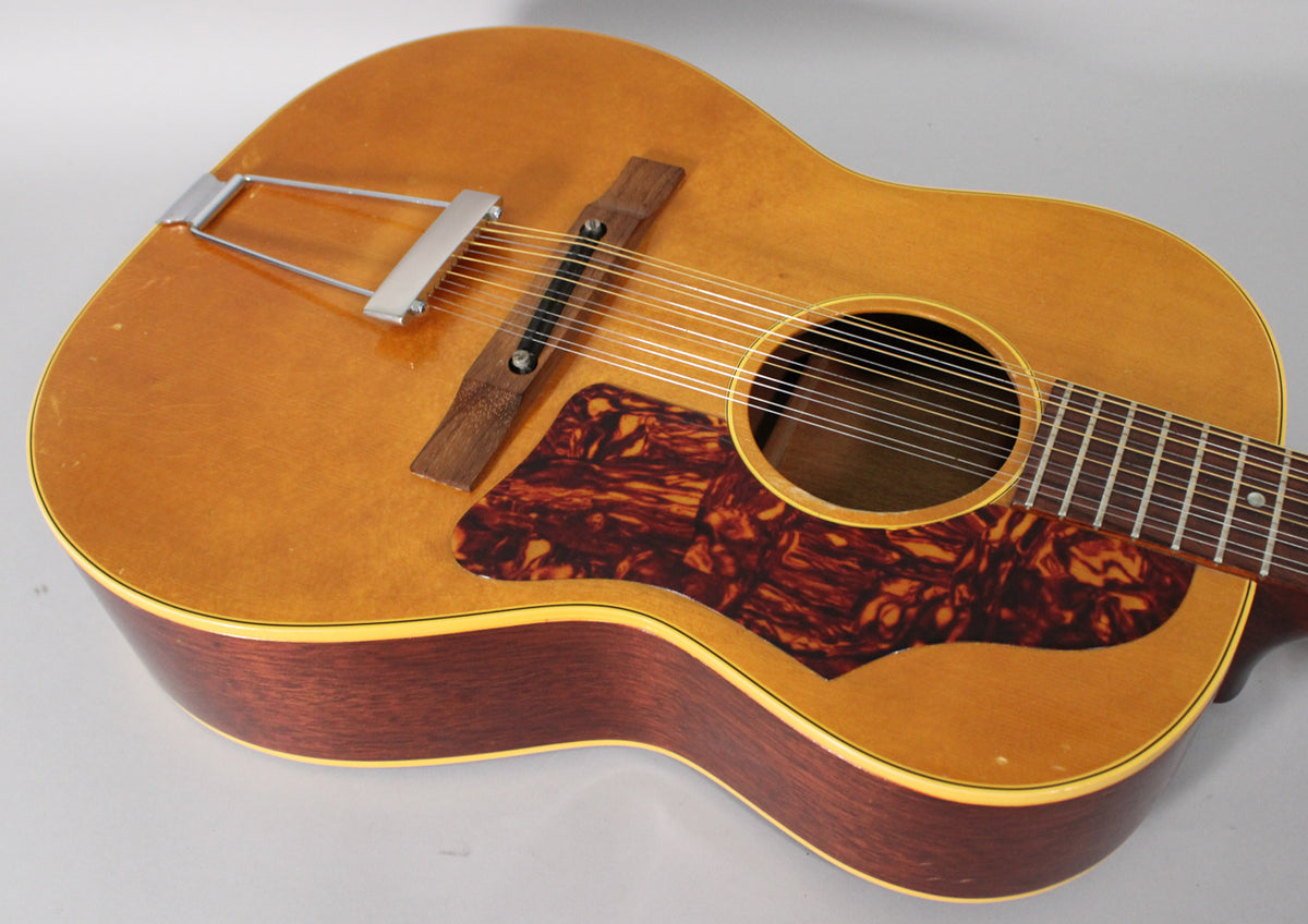 The 12-string acoustic guitar's natural finish top shows light wear, with a tortoiseshell pickguard and a chrome trapeze tailpiece.
