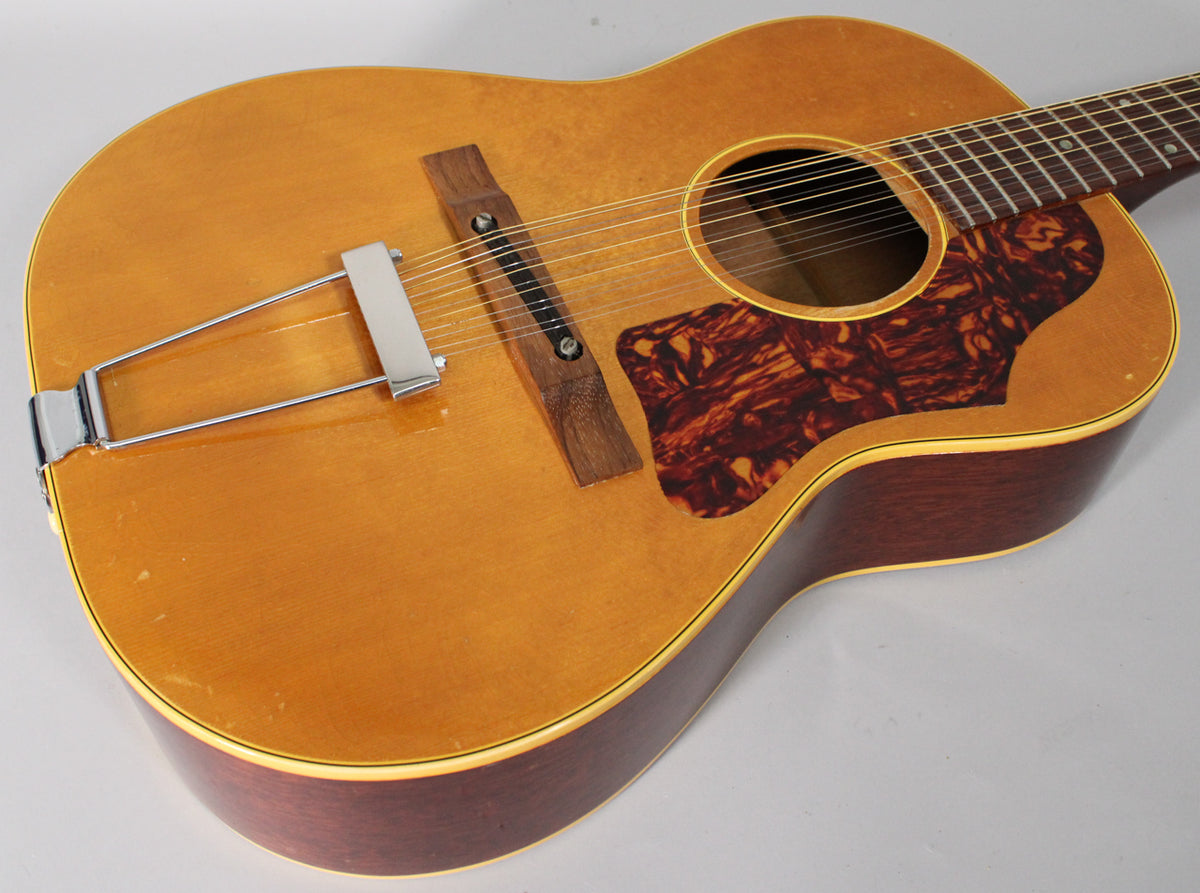 The natural finish 12-string acoustic guitar is angled to show the body, with a tortoiseshell pickguard and chrome trapeze tailpiece.
