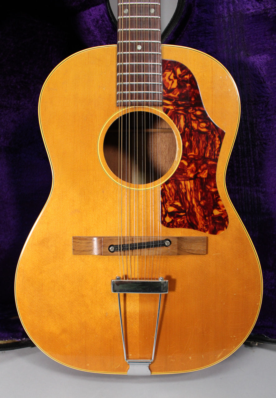 The natural finish 12-string acoustic guitar's body and lower fretboard are visible, showing a tortoiseshell pickguard and chrome trapeze tailpiece.