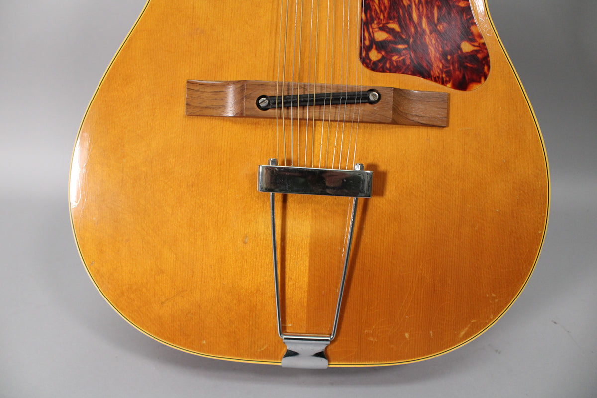 The lower body of the natural finish 12-string acoustic guitar, showing the wooden bridge, chrome trapeze tailpiece, and tortoiseshell pickguard.