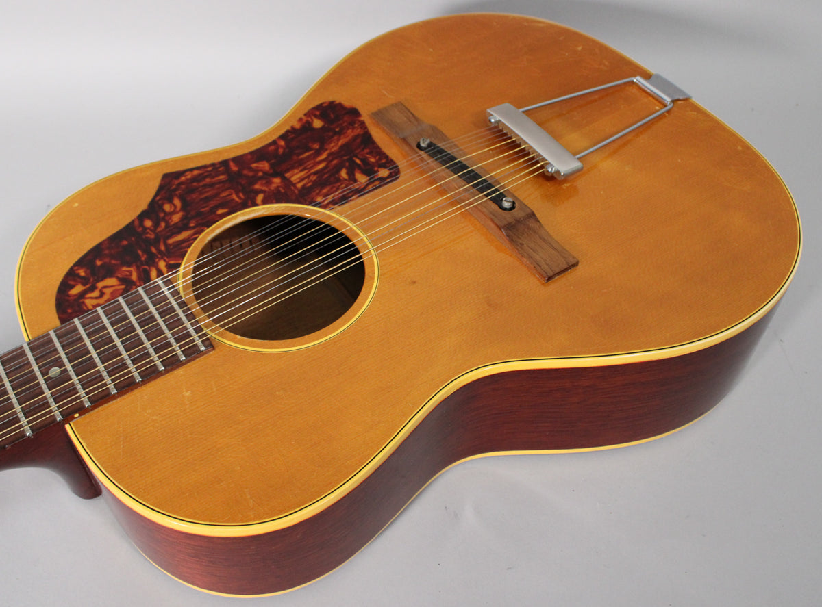 The natural finish 12-string acoustic guitar is angled from the upper left, showing its tortoise pickguard, rosewood bridge, and trapeze tailpiece.