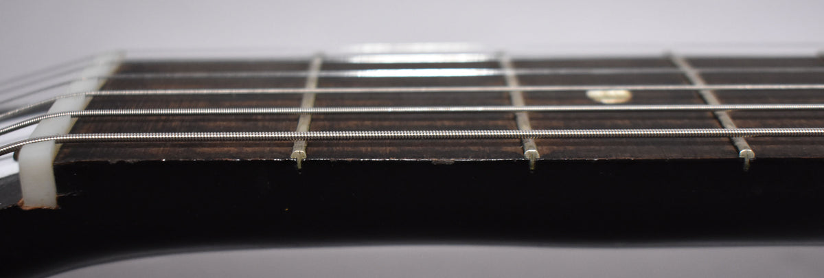 Close-up of the dark fretboard and frets, showing a white nut, light strings, and a small, round fret marker.