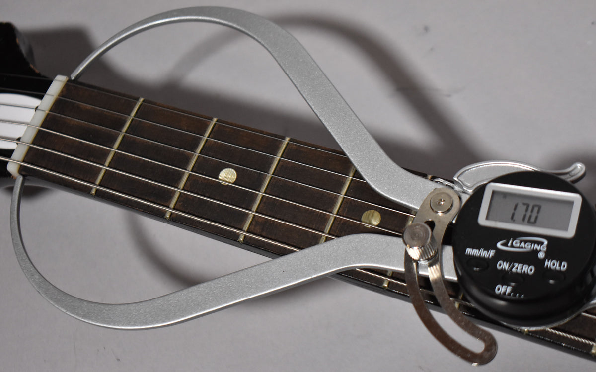 A digital caliper measures the guitar's dark fretboard and strings, showing "1.70" on its display.