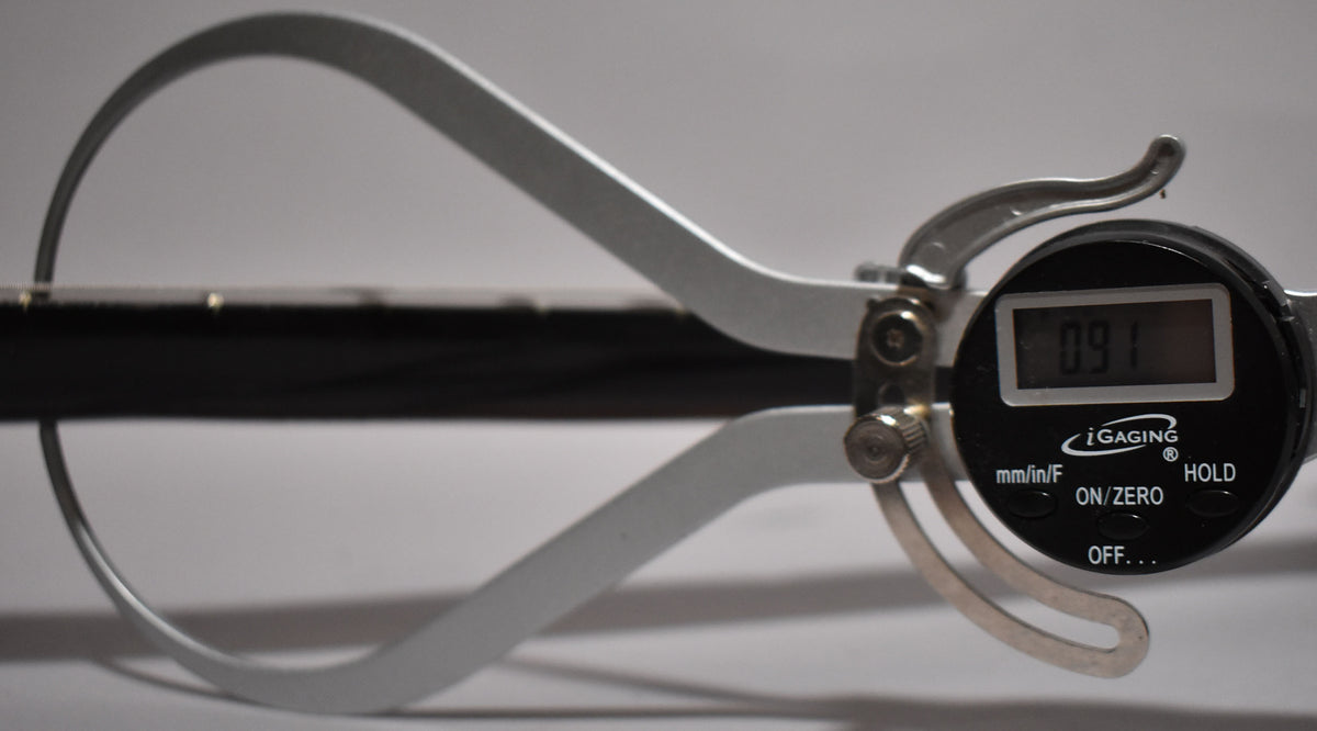 A digital caliper measures the black guitar neck, showing a reading of 0.91, with the fretboard and small dot inlays visible.