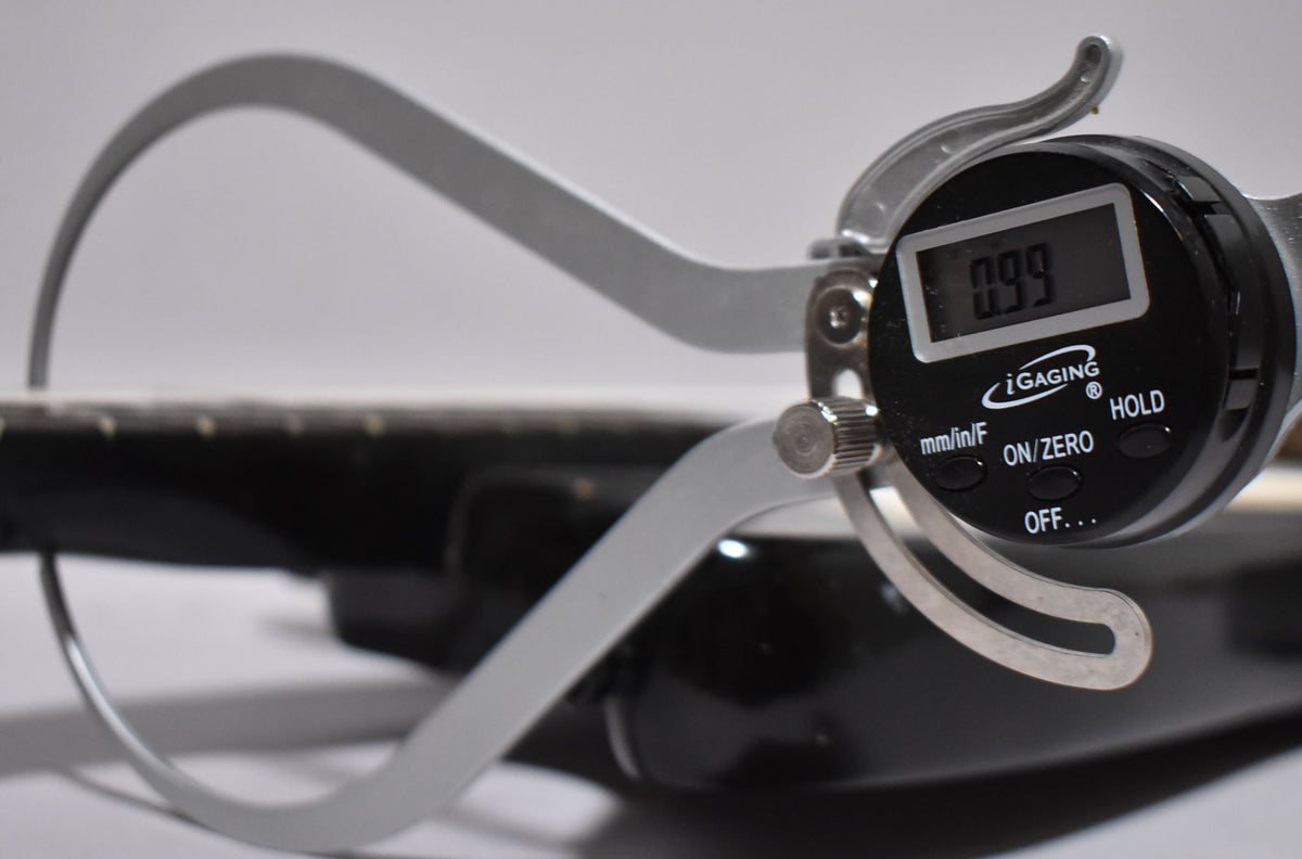 A digital caliper measures the black guitar's fretboard, displaying "0.99" on its screen.