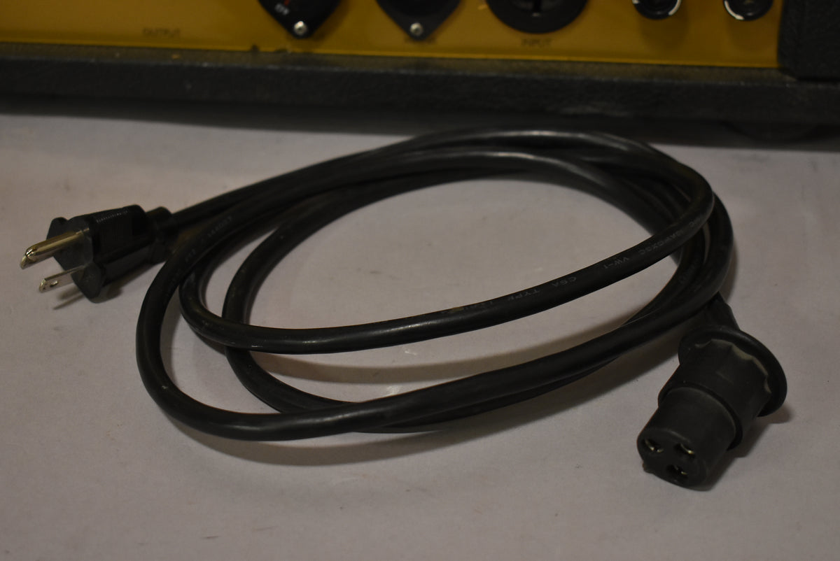 A coiled black power cable with a standard US plug and a 3-pin IEC C13 connector, partially obscuring the amp's yellow rear panel.