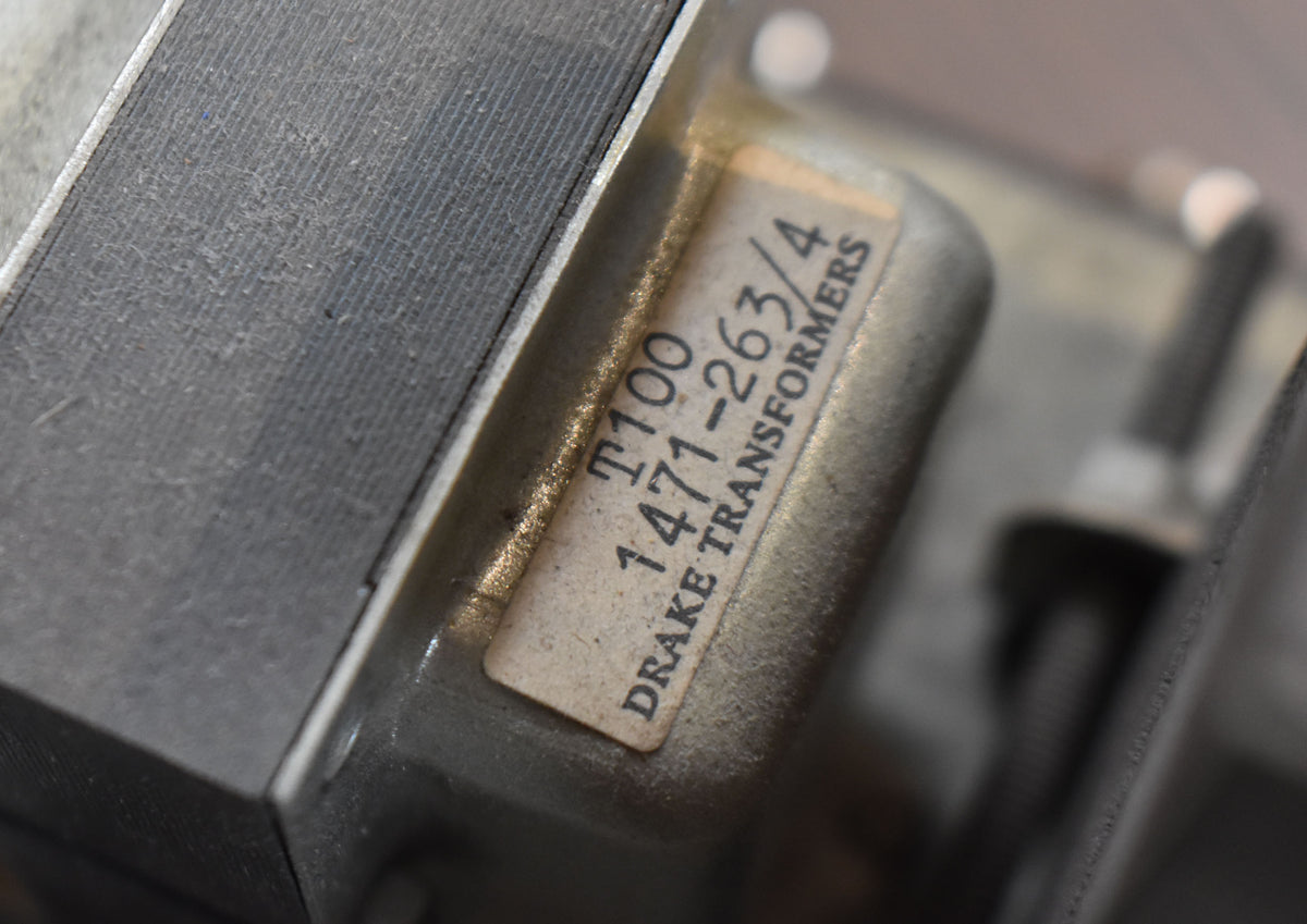 Close-up of a silver-grey transformer with a faded white label showing "T100 1471-263/4 DRAKE TRANSFORMERS".