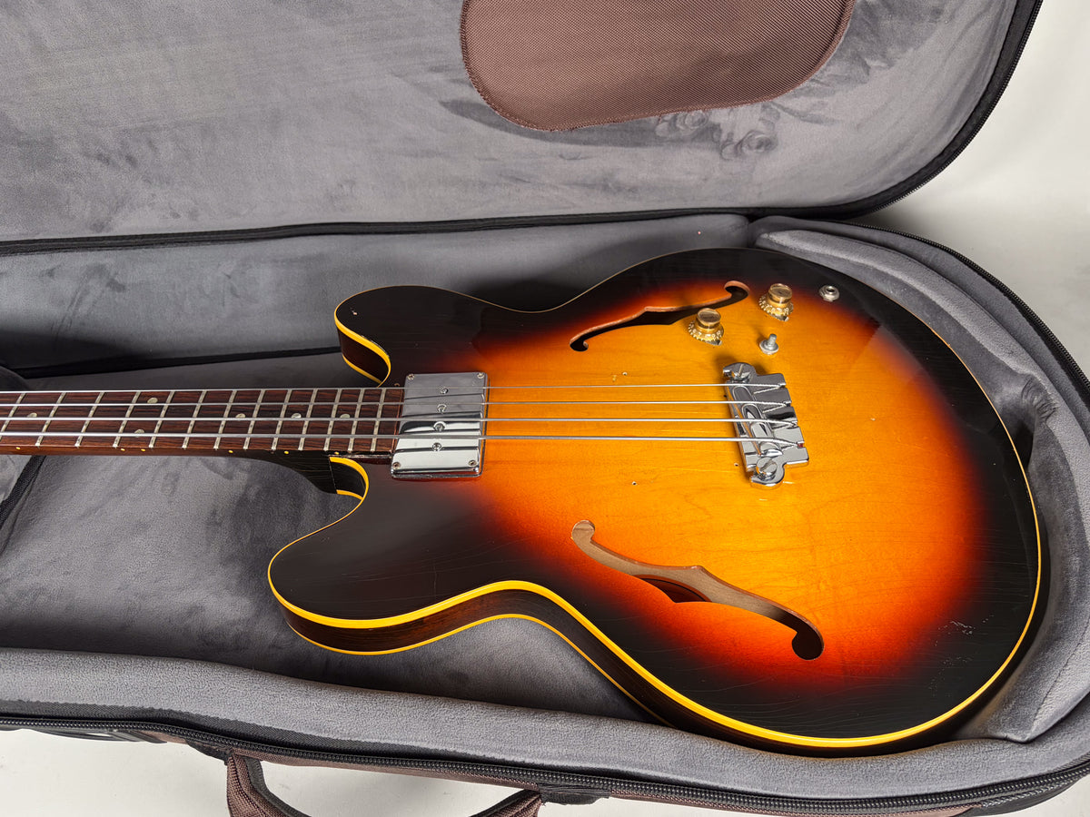 The sunburst hollow-body bass lies in its soft case, showing its f-holes, chrome pickup, bridge, and four gold control knobs.