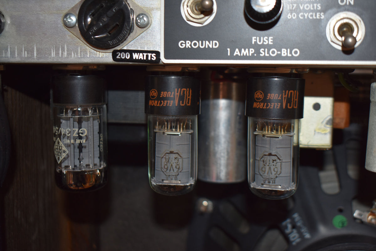 Close-up of three glowing vacuum tubes, two with "RCA" branding, mounted below a silver control panel with a fuse and power switch.