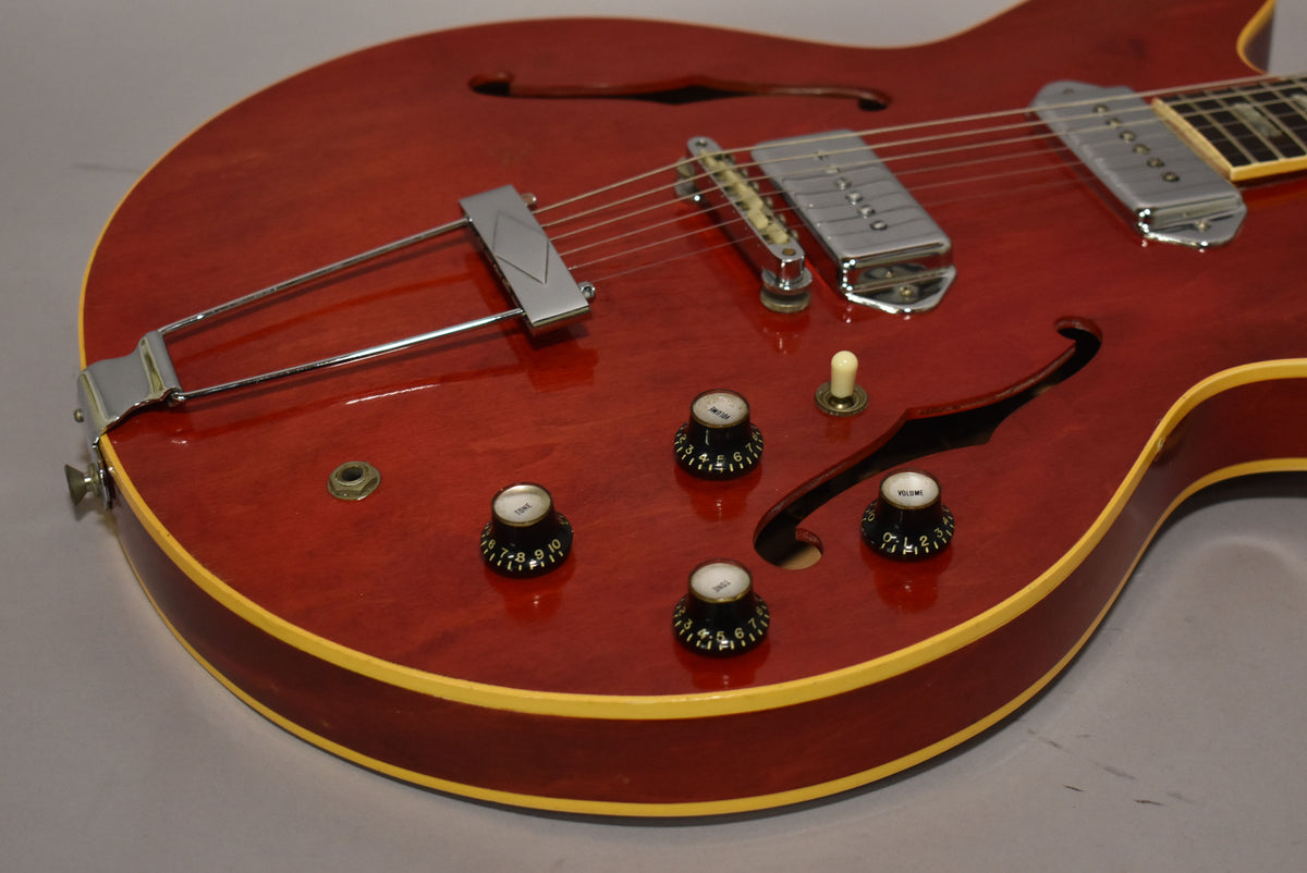 Close-up of the cherry red hollowbody's lower bout, showing the trapeze tailpiece, two chrome P90 pickups, and four black control knobs.