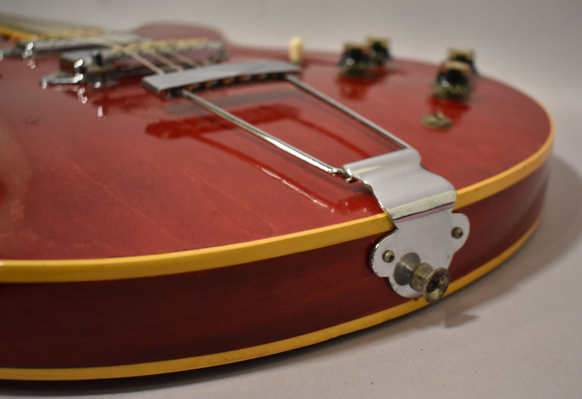 Close-up of the cherry red hollowbody guitar's lower bout, showing the chrome trapeze tailpiece, output jack, and body binding.