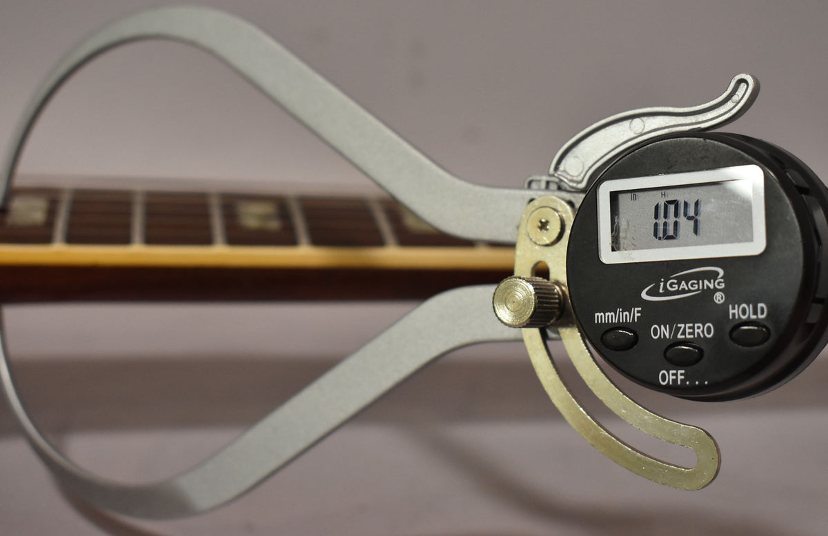 A digital caliper measures the guitar's neck, showing "1.04" on its screen, with the fretboard visible in the background.