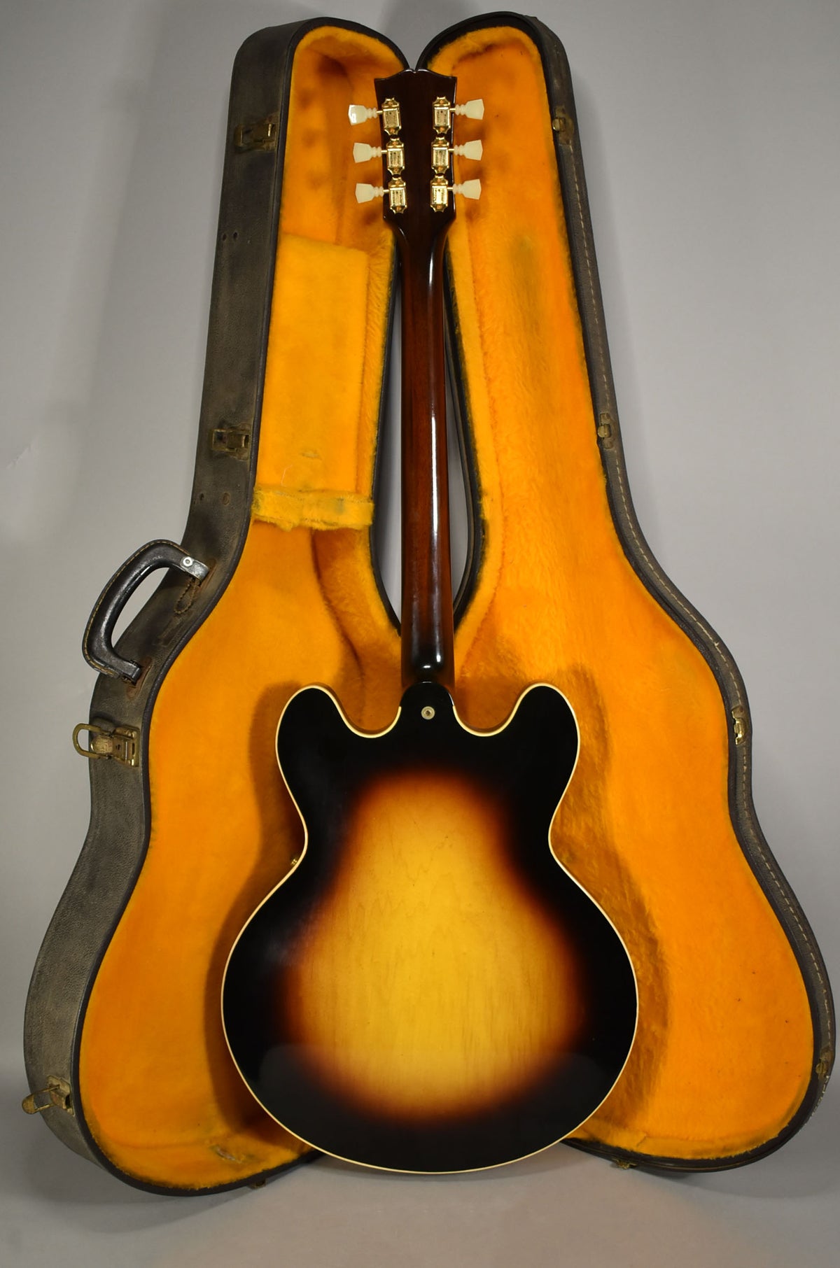 The guitar's sunburst back and neck are visible, with six gold tuners and cream buttons, resting in an open, orange-lined hard case.