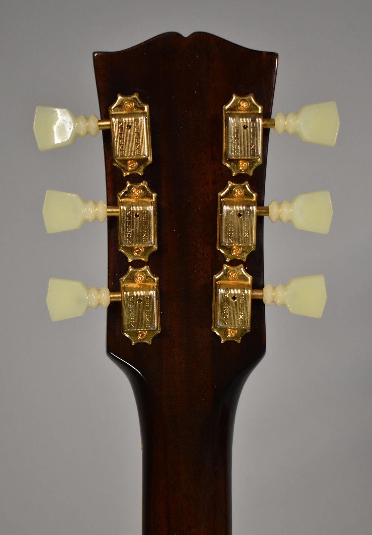 The back of a dark brown headstock with six gold tuners and cream plastic buttons, showing light wear.