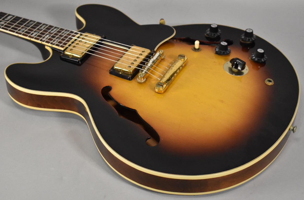 The sunburst semi-hollowbody guitar, angled left, shows gold hardware, black knobs, and a cream binding with some finish wear.