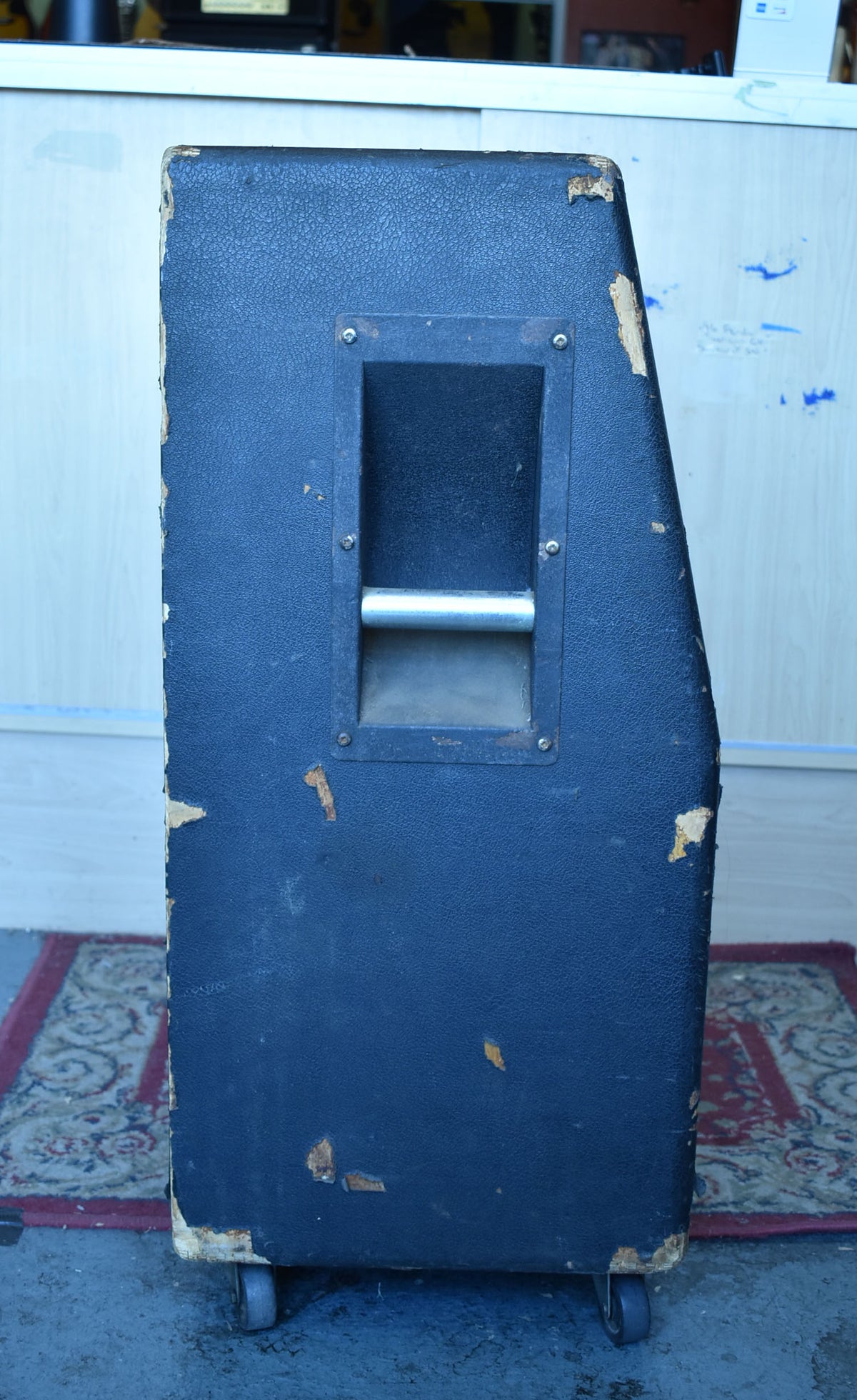 The side of a black basketweave cabinet, showing a recessed metal handle, worn tolex, and a bottom caster wheel.