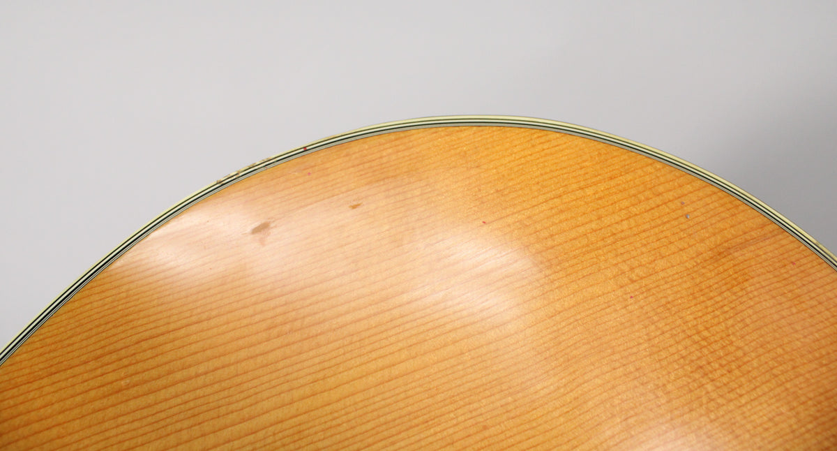 Close-up of the guitar's light-colored, fine-grained spruce top, showing a few small dark spots and a thin black and white binding.
