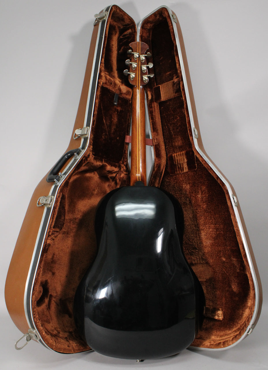 The guitar's shiny black bowl back and brown neck are seen from behind, resting in an open brown hard case with plush lining.